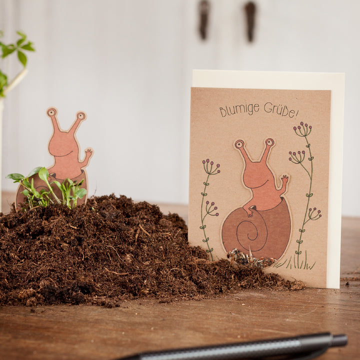 Tarjeta de felicitación con motivo de caracol y la inscripción "saludos florales" junto a un montón de tierra sobre una mesa de madera.