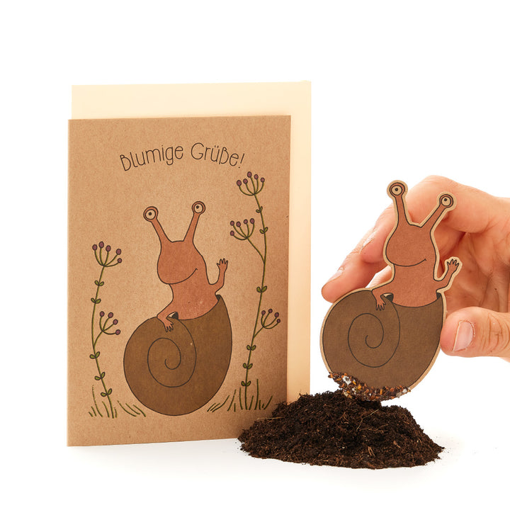 Persona que sostiene una ilustración de papel recortada de un caracol sobre tierra real, a juego con una tarjeta de felicitación con el texto "¡Saludos floridos!" y un dibujo de un caracol.