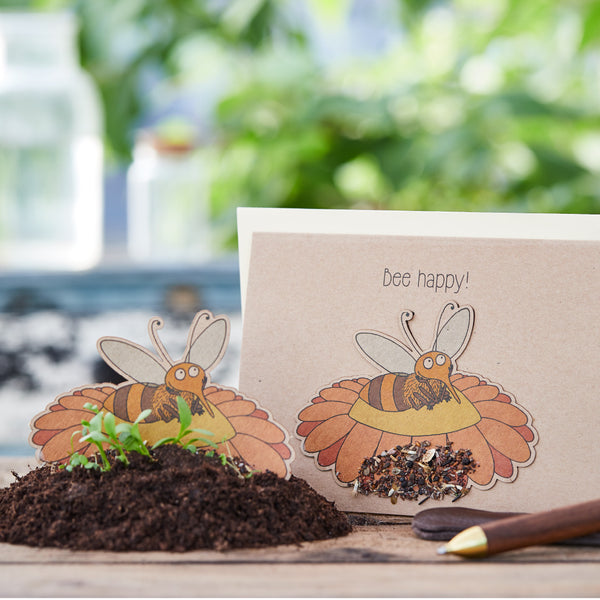 Manualidad con motivo de abeja y la inscripción "¡Abeja feliz!" sobre una mesa con tierra y plantas de fondo.
