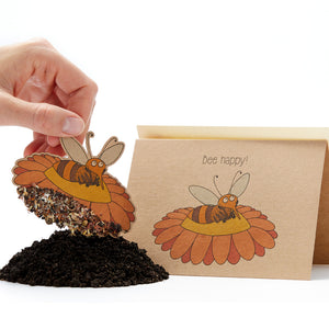 Una mano que sostiene una tarjeta de semillas con forma de abeja con el lema "¡Abeja feliz!" y un montón de tierra debajo.