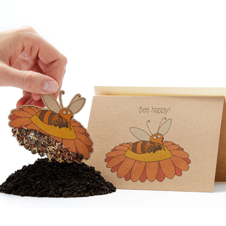Una mano que sostiene una tarjeta de semillas con forma de abeja con el lema "¡Abeja feliz!" y un montón de tierra debajo.