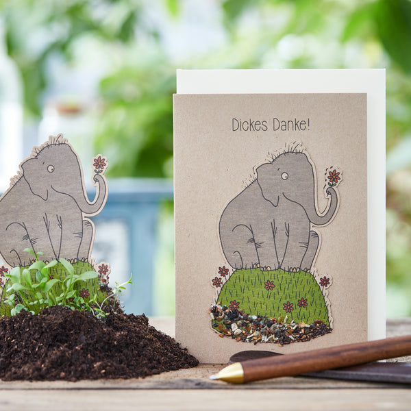 Tarjeta de felicitación con un mamut dibujado que dice “¡Muchas gracias!”, junto a él tierra y plántulas verdes.