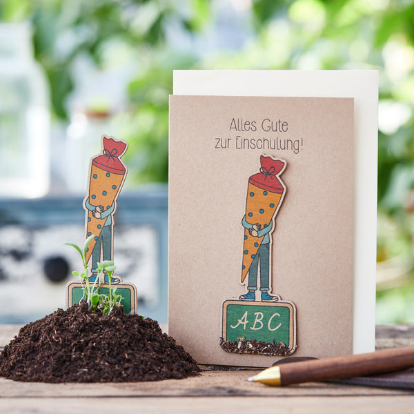 Tarjeta de felicitación por inscripción escolar con figura de zanahoria y tablero ABC sobre una mesa de madera, rodeada de plantas y tierra.