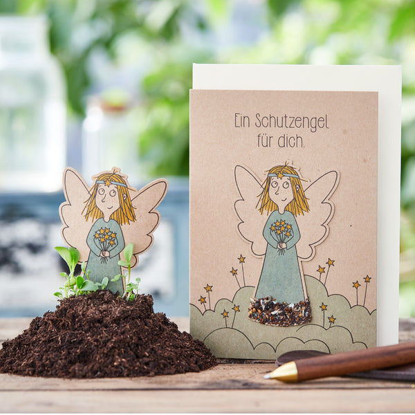 Tarjeta de felicitación con motivo de ángel de la guarda junto a montículo de tierra y bolígrafo sobre mesa de madera.