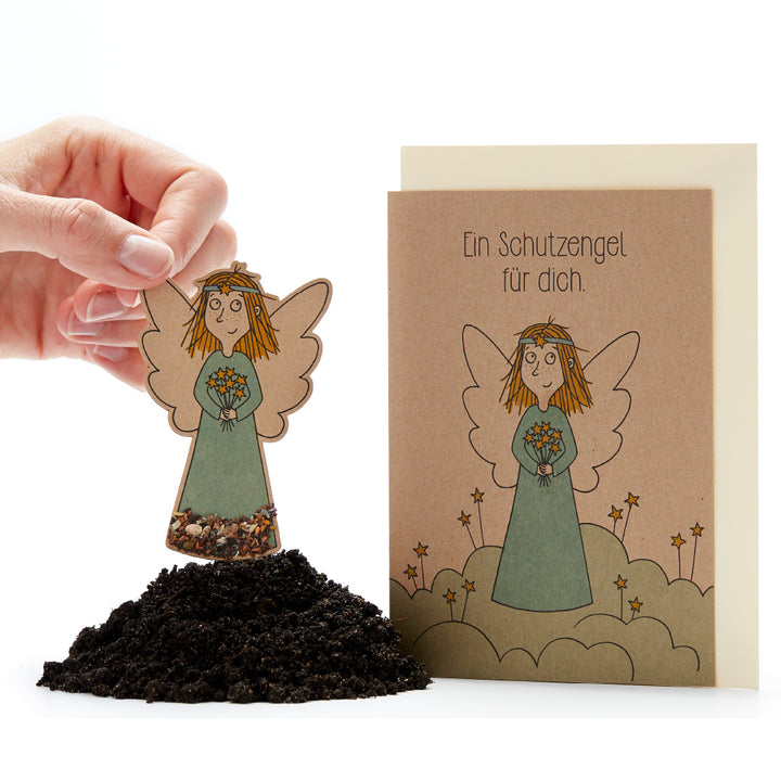 Mano sosteniendo una figura de ángel hecha de papel con semillas sobre tierra junto a una tarjeta con el texto 'Un ángel de la guarda para ti'.
