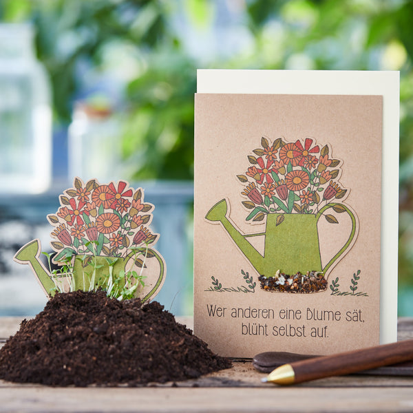 Tarjeta de felicitación con la ilustración de una regadera llena de flores y el dicho alemán "Quien siembra una flor para otros, él mismo florecerá", junto a ella una tarjeta de siembra en una maceta con tierra y vegetación en ciernes.