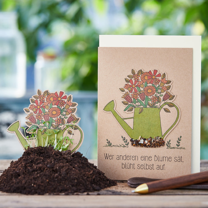 Tarjeta de felicitación con la ilustración de una regadera llena de flores y el dicho alemán "Quien siembra una flor para otros, él mismo florecerá", junto a ella una tarjeta de siembra en una maceta con tierra y vegetación en ciernes.