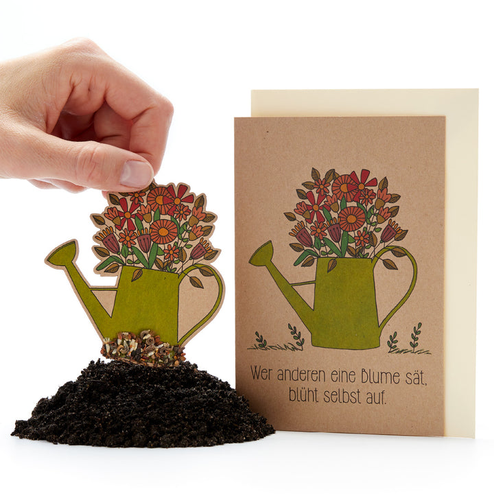 Una mano coloca en la tierra una regadera de papel decorada con flores; junto a ella hay una tarjeta con el mismo motivo y el dicho alemán: "Quien siembra una flor para otros, él mismo florecerá".