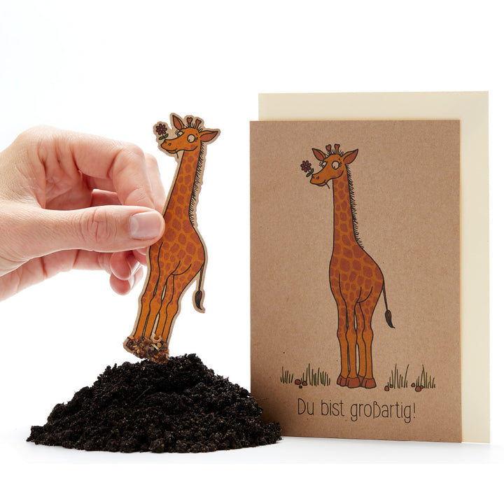 Una mano clava en la tierra una tarjeta con forma de jirafa, junto a ella una tarjeta de felicitación con un dibujo de una jirafa y el texto "¡Eres increíble!".