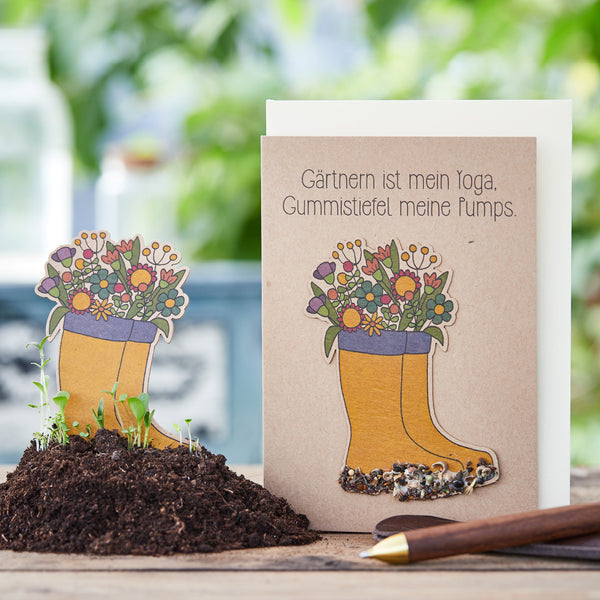 Tarjeta de felicitación con el lema "La jardinería es mi yoga, las botas de goma son mis zapatos" y botas de goma ilustradas llenas de flores sobre un fondo de utensilios de jardinería.