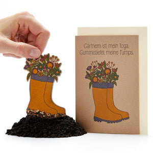 Un arreglo floral en forma de botas de goma, al lado una tarjeta con la inscripción "La jardinería es mi yoga, las botas de goma son mis zapatos".