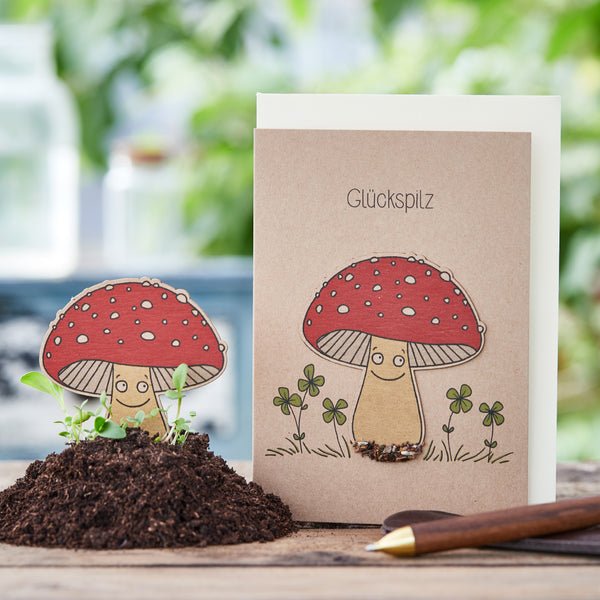 Tarjeta de felicitación con dibujos de hongos y la palabra "hongo de la suerte", junto a un montículo de tierra real y un lápiz.