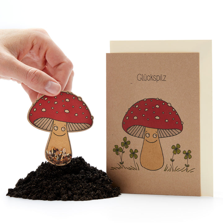 La mano coloca en el suelo una tarjeta con forma de hongo con semillas, junto a ella una tarjeta de felicitación con una amanita muscaria y la inscripción "Hongo de la suerte".