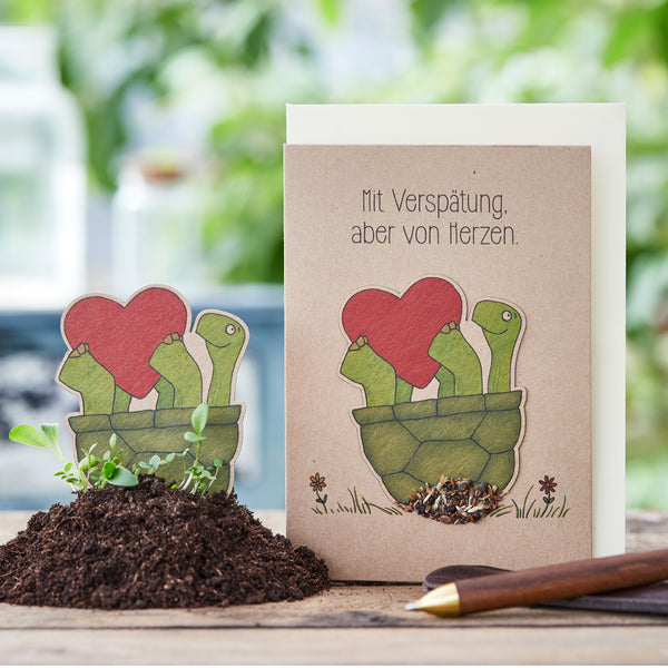 Tarjeta de felicitación con un dibujo de tortugas sosteniendo un corazón rojo, con un marcador de planta a juego en la tierra al lado; el texto en la tarjeta dice: "Retrasado, pero desde el corazón".