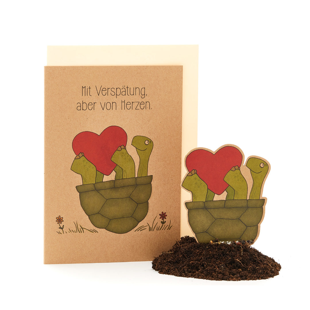 Tarjeta de felicitación con el texto "Tarde, pero de corazón" y una ilustración de plantas parecidas a cactus en una maceta con flores en forma de corazón.