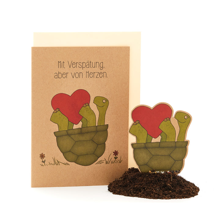 Tarjeta de felicitación con el texto "Tarde, pero de corazón" y una ilustración de plantas parecidas a cactus en una maceta con flores en forma de corazón.