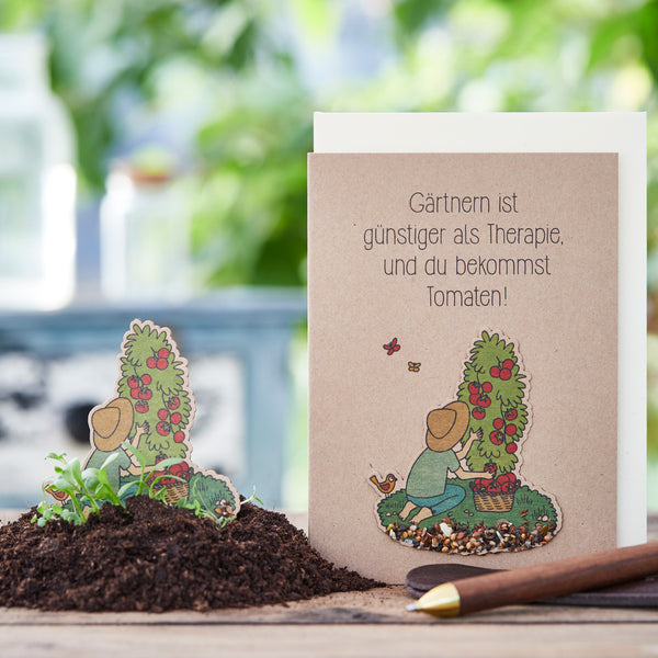 Una tarjeta de felicitación con el lema "La jardinería es más barata que la terapia, ¡y obtienes tomates!" junto a un montón de tierra y una paleta de jardinería.