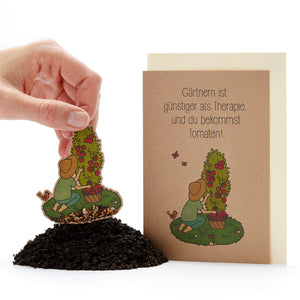 Una mano planta una tarjeta con semillas que representa a un jardinero y una planta de tomate en la tierra, y junto a ella hay una tarjeta de felicitación que dice: "¡La jardinería es más barata que la terapia y obtienes tomates!"