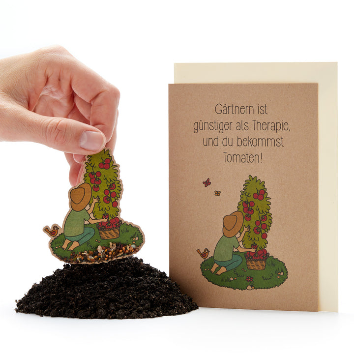 Una mano planta una tarjeta con semillas que representa a un jardinero y una planta de tomate en la tierra, y junto a ella hay una tarjeta de felicitación que dice: "¡La jardinería es más barata que la terapia y obtienes tomates!"