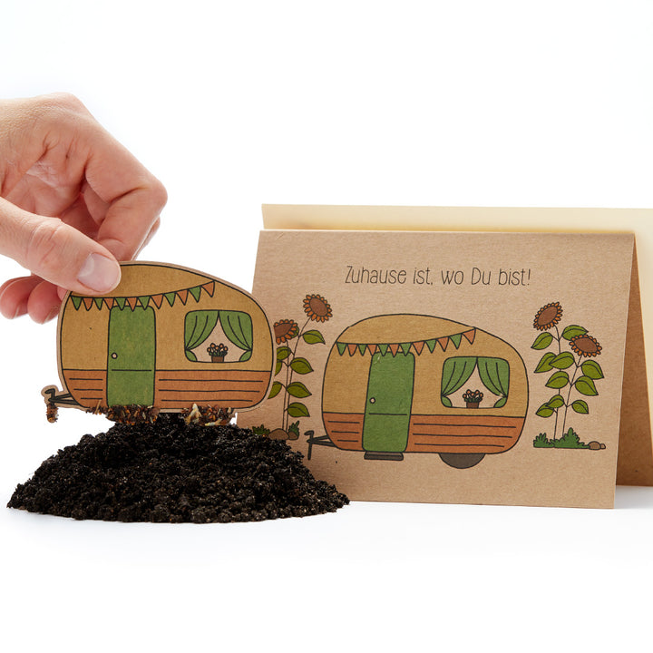 Mano colocando una tarjeta de plantación con una caravana ilustrada en la tierra. El texto "¡Tu hogar está donde estás!" está escrito en la tarjeta.