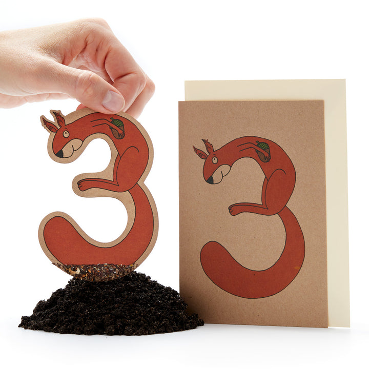 Mano sosteniendo una figura de planta de ardilla roja junto a una tarjeta de felicitación con el mismo diseño sobre un fondo blanco.