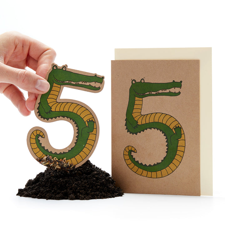 Mano sosteniendo una figura de papel recortada en forma de "5" que parece un cocodrilo, junto a una tarjeta con el mismo diseño en la tierra.