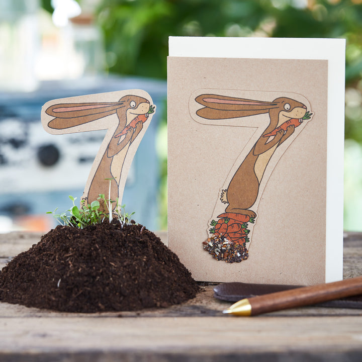 Tarjeta infantil con el número siete, que representa un conejo con una zanahoria, junto a él un macizo de flores con tierra y plantas.