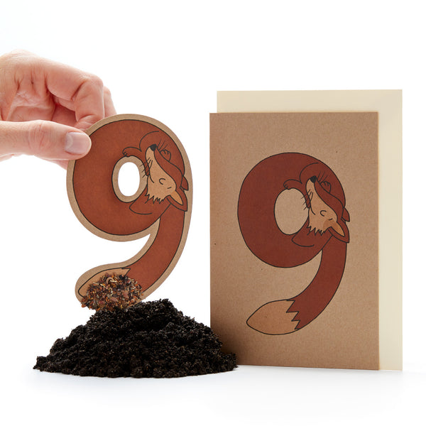 Mano sosteniendo una letra "ö" troquelada con un diseño de zorro junto a una tarjeta con el mismo diseño y un pequeño montón de tierra.