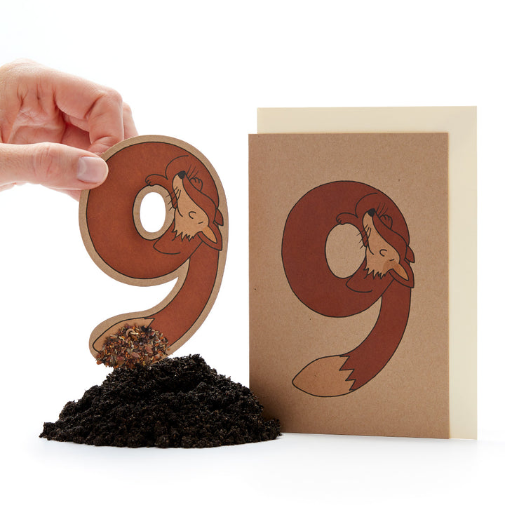Mano sosteniendo una letra "ö" troquelada con un diseño de zorro junto a una tarjeta con el mismo diseño y un pequeño montón de tierra.