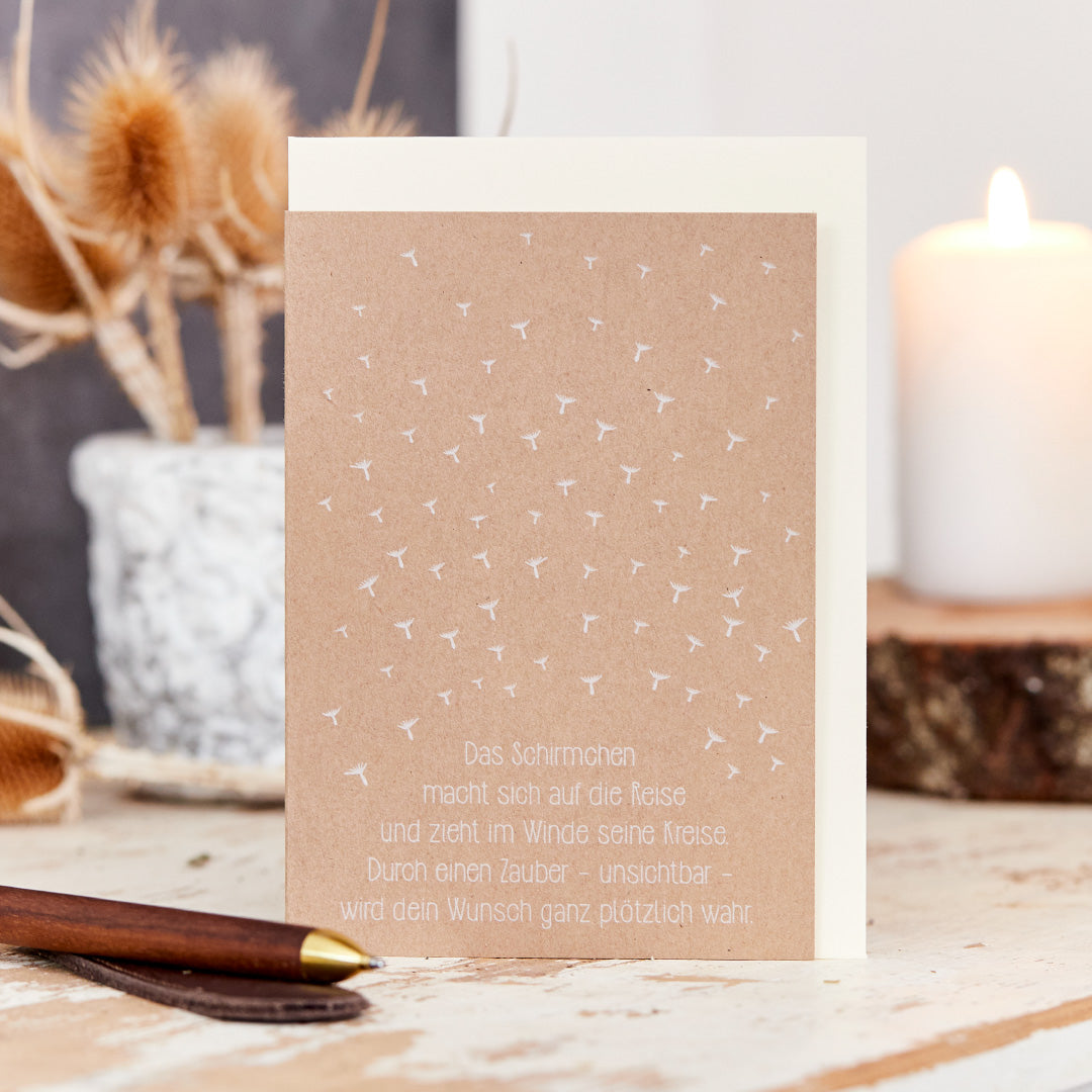 Tarjeta de felicitación con motivo de paraguas y dicho alemán, sobre una mesa de madera junto a flores secas y una vela.