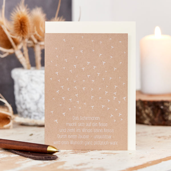 Tarjeta de felicitación con motivo de paraguas y dicho alemán, sobre una mesa de madera junto a flores secas y una vela.