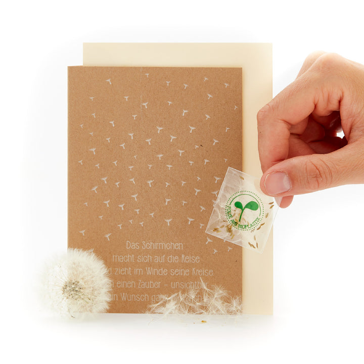 Mano sosteniendo una tarjeta de felicitación con semillas de diente de león y un poema junto a un diente de león.
