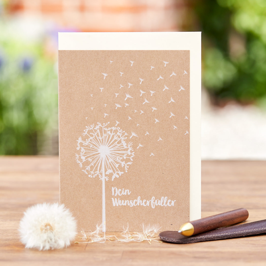 Tarjeta de felicitación con un diente de león y la inscripción "Tu Cumplidor de Deseos", junto a un diente de león real y un bolígrafo sobre un fondo de madera.