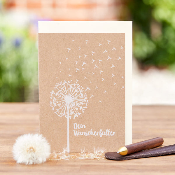Tarjeta de felicitación con un diente de león y la inscripción "Tu Cumplidor de Deseos", junto a un diente de león real y un bolígrafo sobre un fondo de madera.