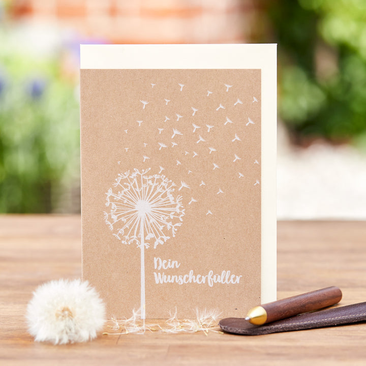 Tarjeta de felicitación con un diente de león y la inscripción "Tu Cumplidor de Deseos", junto a un diente de león real y un bolígrafo sobre un fondo de madera.