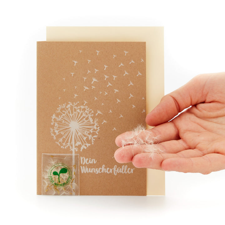 Mano sosteniendo una tarjeta con un diseño de diente de león y la inscripción "Tu Cumplidor de Deseos" y una pequeña bolsa transparente con semillas de diente de león.