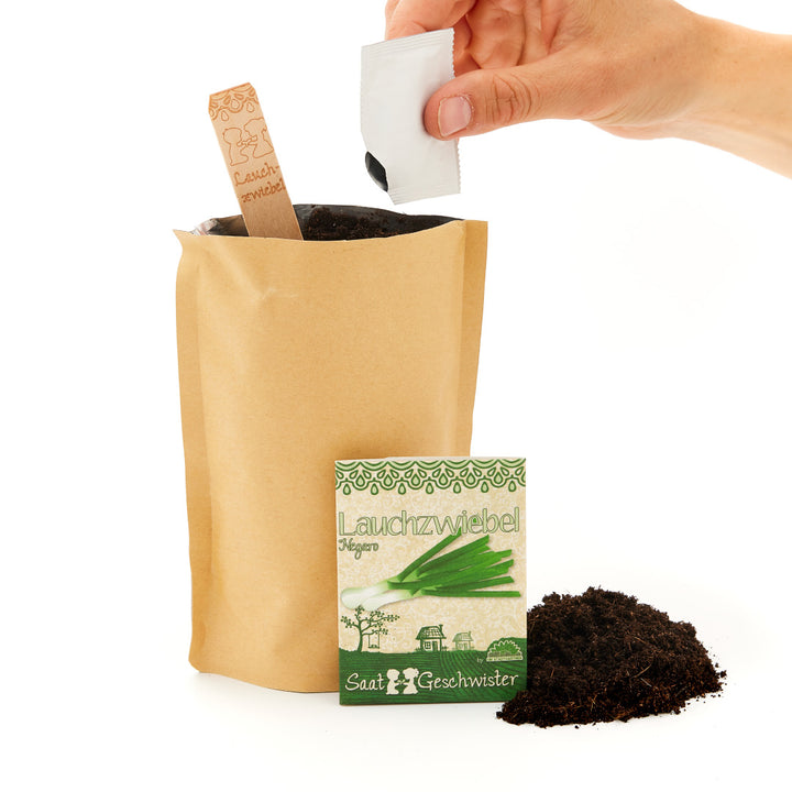 Mano sosteniendo un paquete de semillas sobre una bolsa de papel marrón con tierra, al lado de otro paquete de semillas y una pila de tierra.