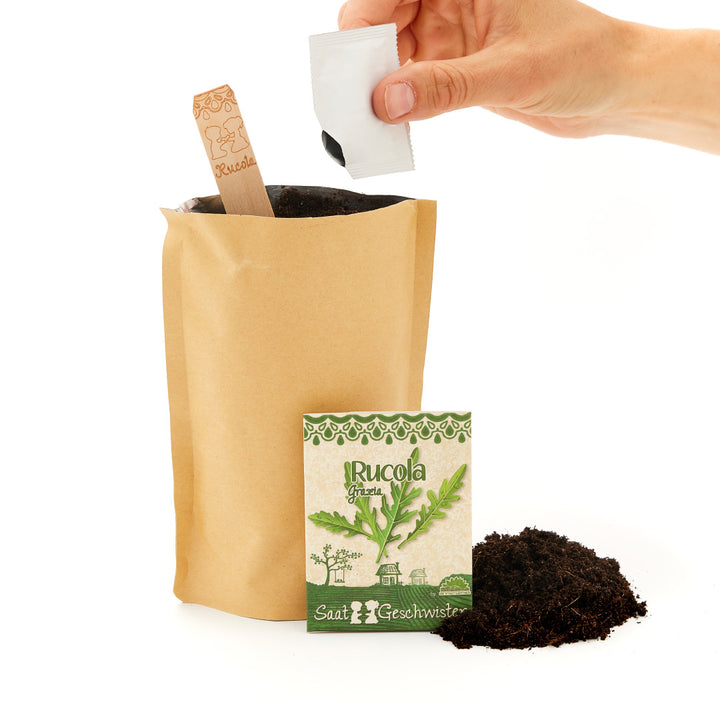 Una mano abre un paquete de semillas de rúcula junto a una bolsa de papel con tierra y un cartel de madera que dice "Rúcula".