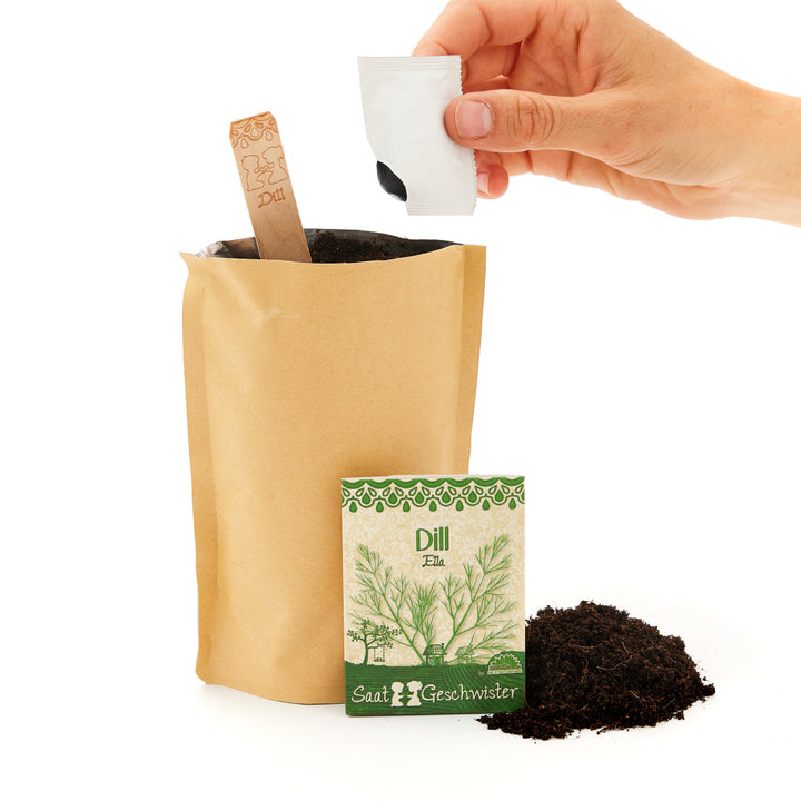 Mano vertiendo semillas en una bolsa de papel para maceta, al lado de un paquete de semillas de eneldo y una pala de tierra.