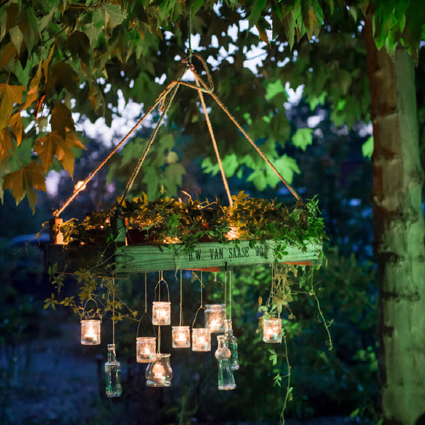 Cesta de flores colgante con plantas y velas en vasos iluminados con la luz del atardecer.