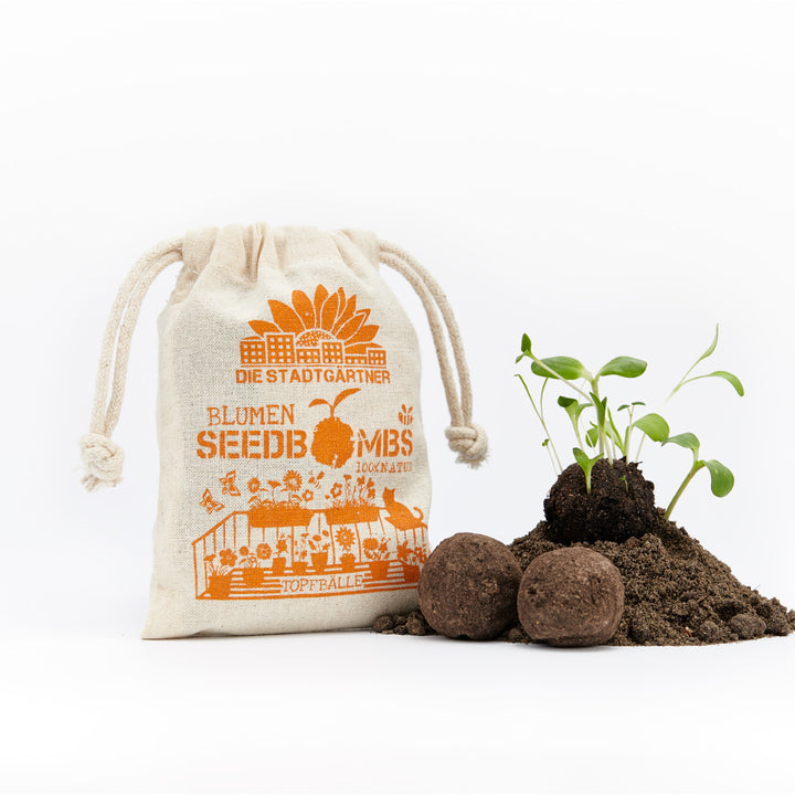 Bolsa de tela con la inscripción "DIE STADTGÄRTNER BLUMEN SEEDBOMBS 100% NATUR" y junto a ella plantas germinando sobre un montículo de tierra con dos bombas de semillas.