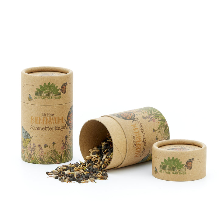 Mezcla de semillas para abejas y mariposas en dos tubos de cartón con la inscripción 'Aktion BIENENWOHL Schmetterlinge' y contenido derramado junto a un tubo volcado.