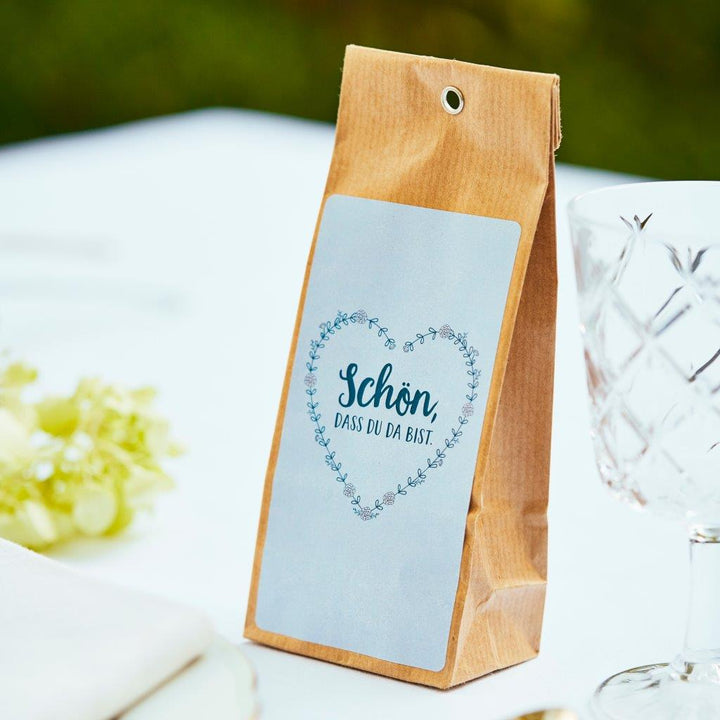 Bolsa de papel marrón con la inscripción "Es agradable tenerte aquí" sobre una mesa junto a una copa de vino y uvas.