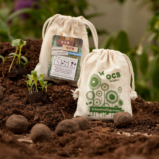 Dos bolsas de tela etiquetadas "OCB Organic" con crecimiento de plantas jóvenes sobre una capa de tierra, con un paisaje de jardín en el fondo.